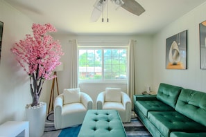 A living room with a green sofa, two white armchairs, a green ottoman, a large window with curtains, a ceiling fan, and a decorative pink tree.