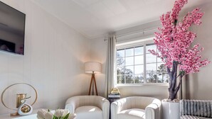 Ein Wohnzimmer mit zwei weißen Ledersofas, einem Couchtisch mit einer Vase weißer Blumen, einer Stehlampe und einem großen Fenster mit Blick auf Bäume.