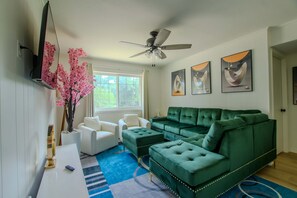 Ein Wohnzimmer mit einem grünen Ecksofa, weißen Sessel, einem Flachbildfernseher, einem Deckenventilator und Kunstwerken an der Wand.