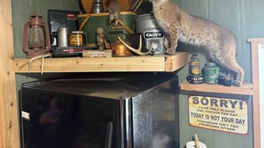 A cat on a shelf with camping gear, a sign saying "SORRY! I can only please one person per day," and a refrigerator.