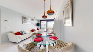 Un salon moderne avec un canapé blanc, des coussins rouges et bleus, une table à manger en verre avec des assiettes rouges, des chaises à motifs et une grande fenêtre donnant sur des immeubles.