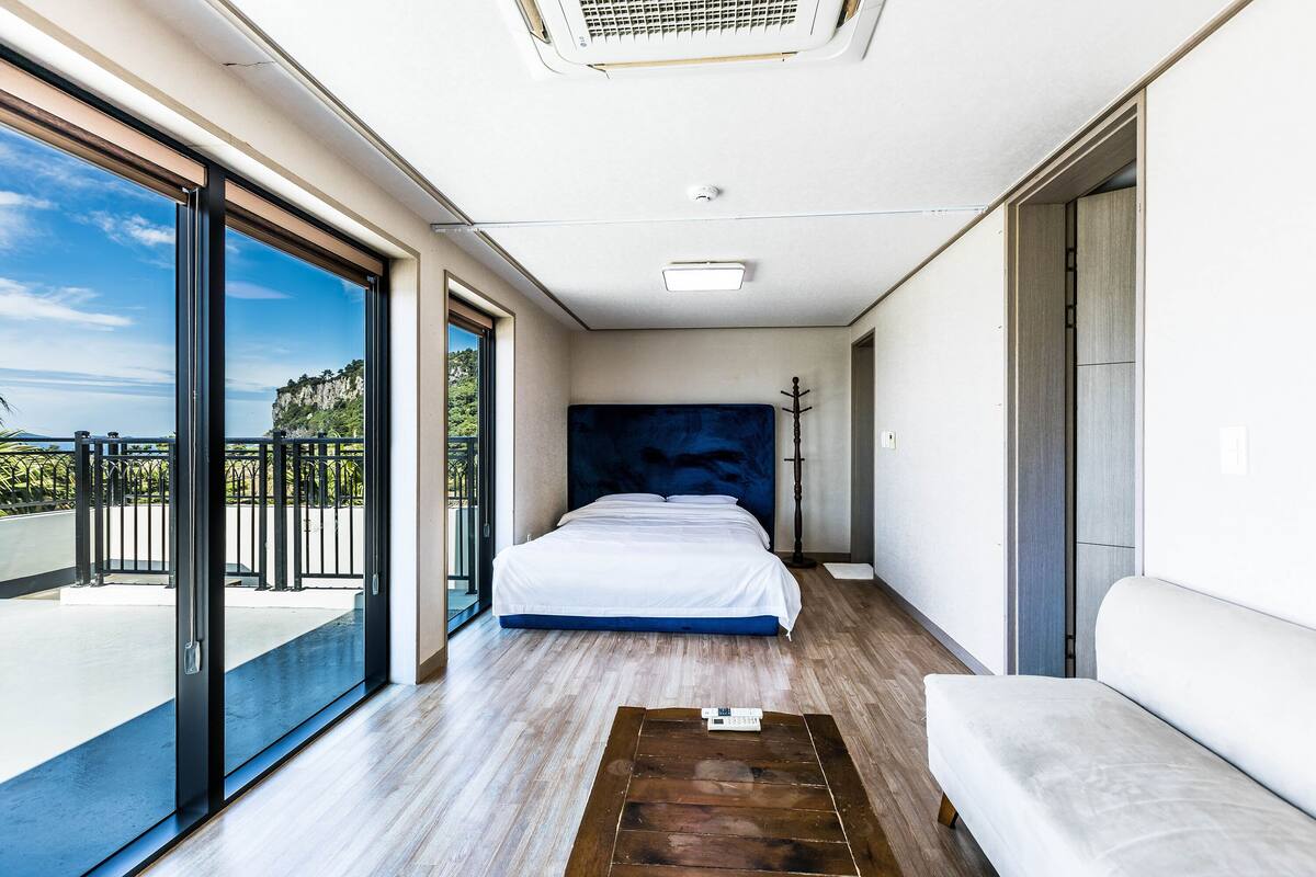 A modern bedroom with a large bed, a wooden floor, a white sofa, and a balcony with a view of the sea.