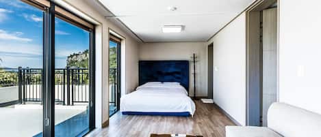 A modern bedroom with a large bed, a wooden floor, a white sofa, and a balcony with a view of the sea.
