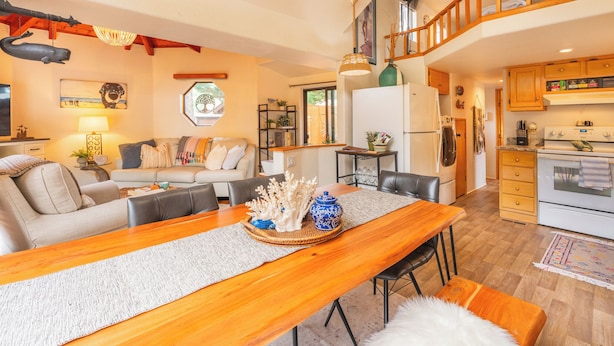 Un salon moderne avec une table à manger en bois, un ensemble de canapés et un coin cuisine doté de meubles en bois et d’un four encastré.