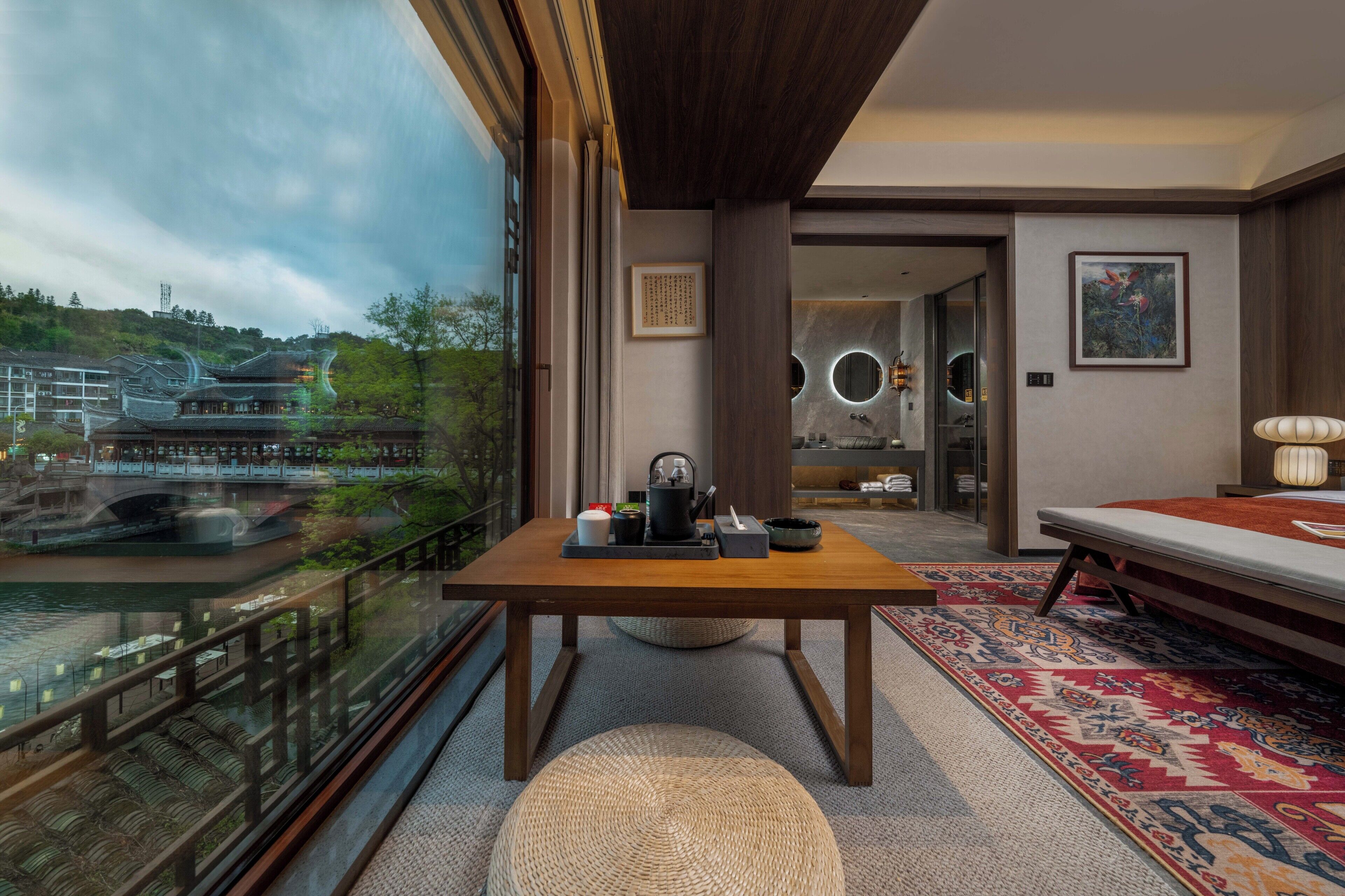 A modern hotel room with a wooden table, a sofa, a view of a traditional building, and a patterned rug.