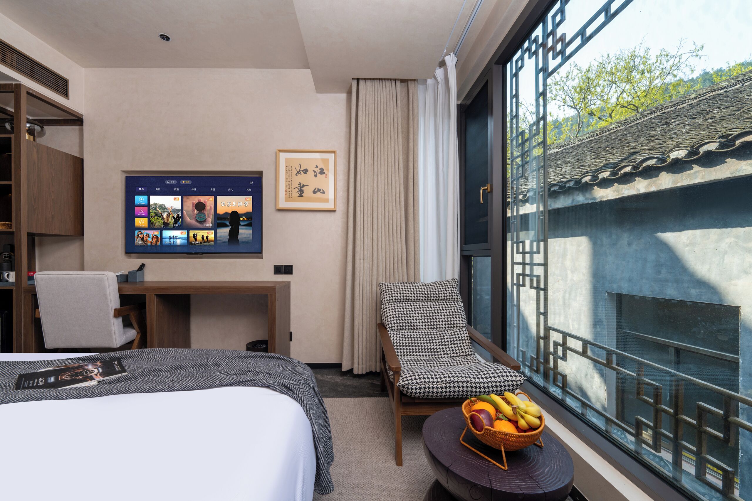 A hotel room with a bed, a desk, a chair, a TV displaying photos, a framed artwork, and a window with a view of a building and trees.