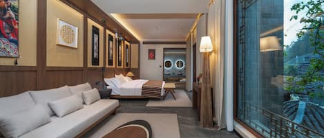 Ein modernes Hotelzimmer mit einer Couch, einem Bett, einem Holztisch und einem großen Fenster mit Blick auf traditionelle Architektur.