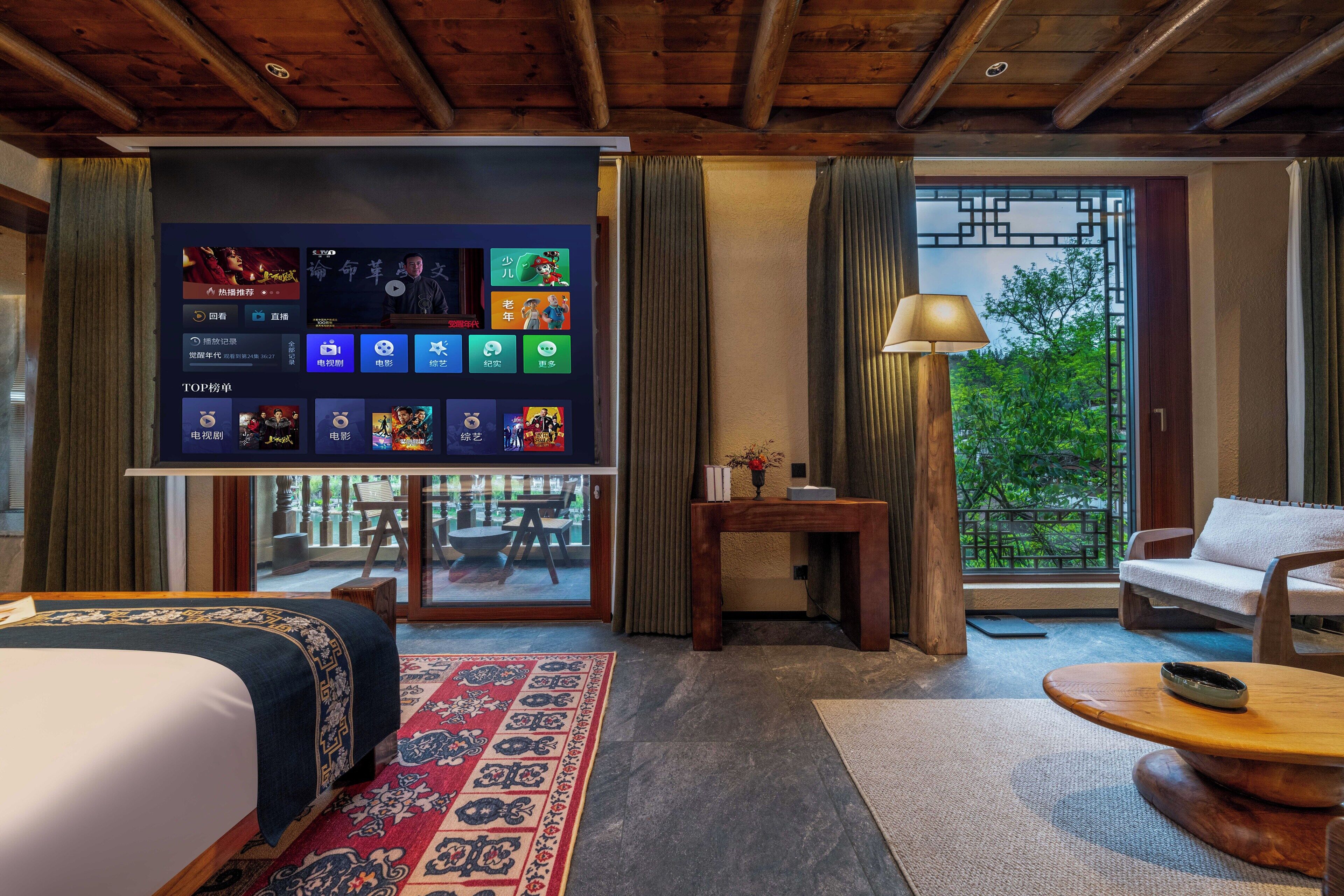 Habitación de hotel con una tele de gran tamaño mostrando diversas aplicaciones, una cama con colcha estampada, una mesita de madera, un sofá y vistas a zonas verdes por la ventana.