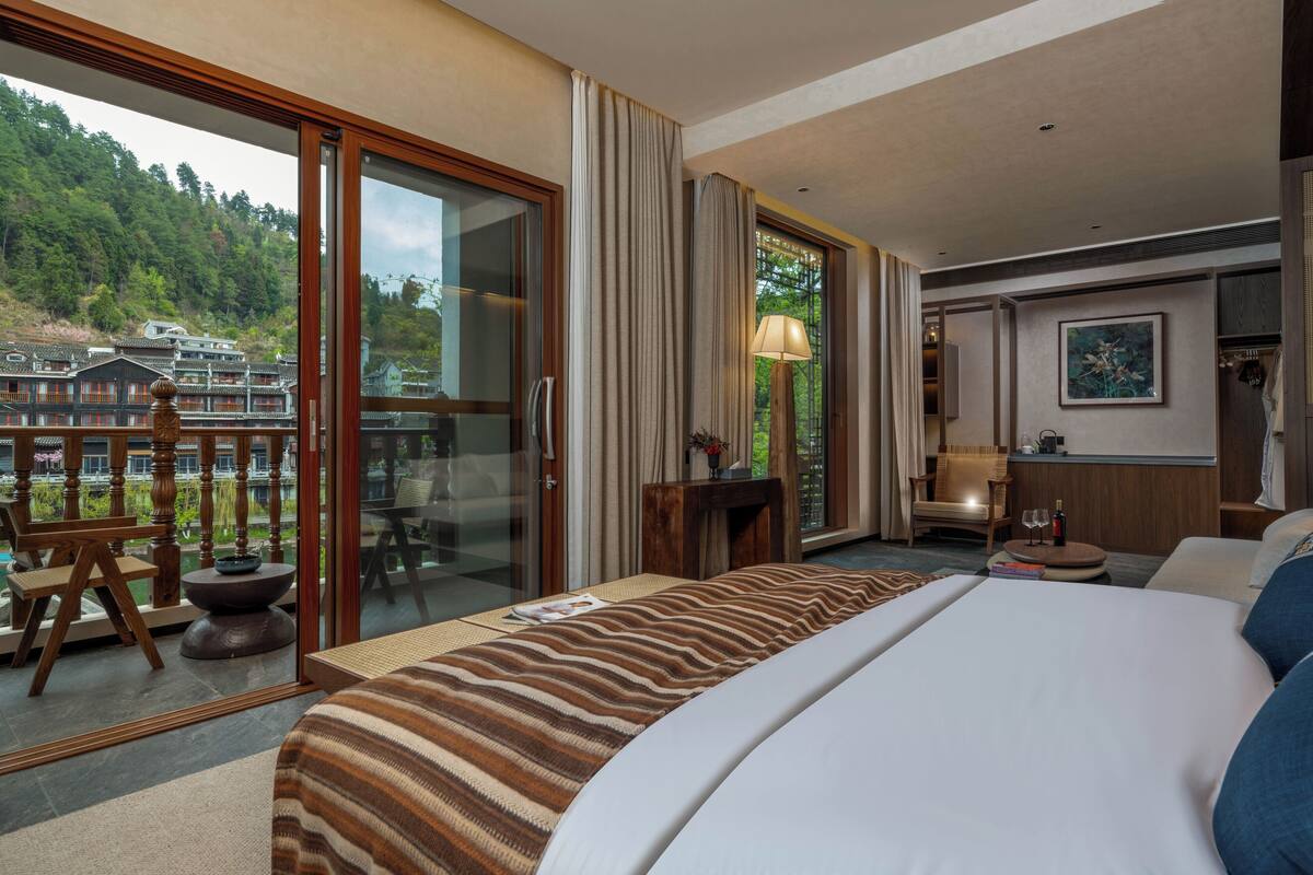 Habitación de hotel con una cama grande, un balcón con vistas a edificios tradicionales, una silla de madera y una mesita con una lámpara.