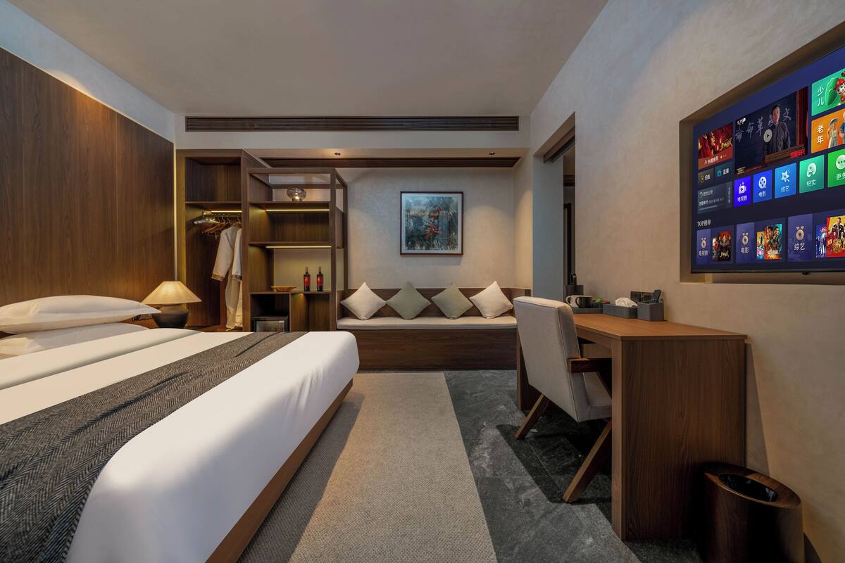Habitación de hotel con cama, escritorio, zona de estar, armario y televisor empotrado mostrando varios canales.