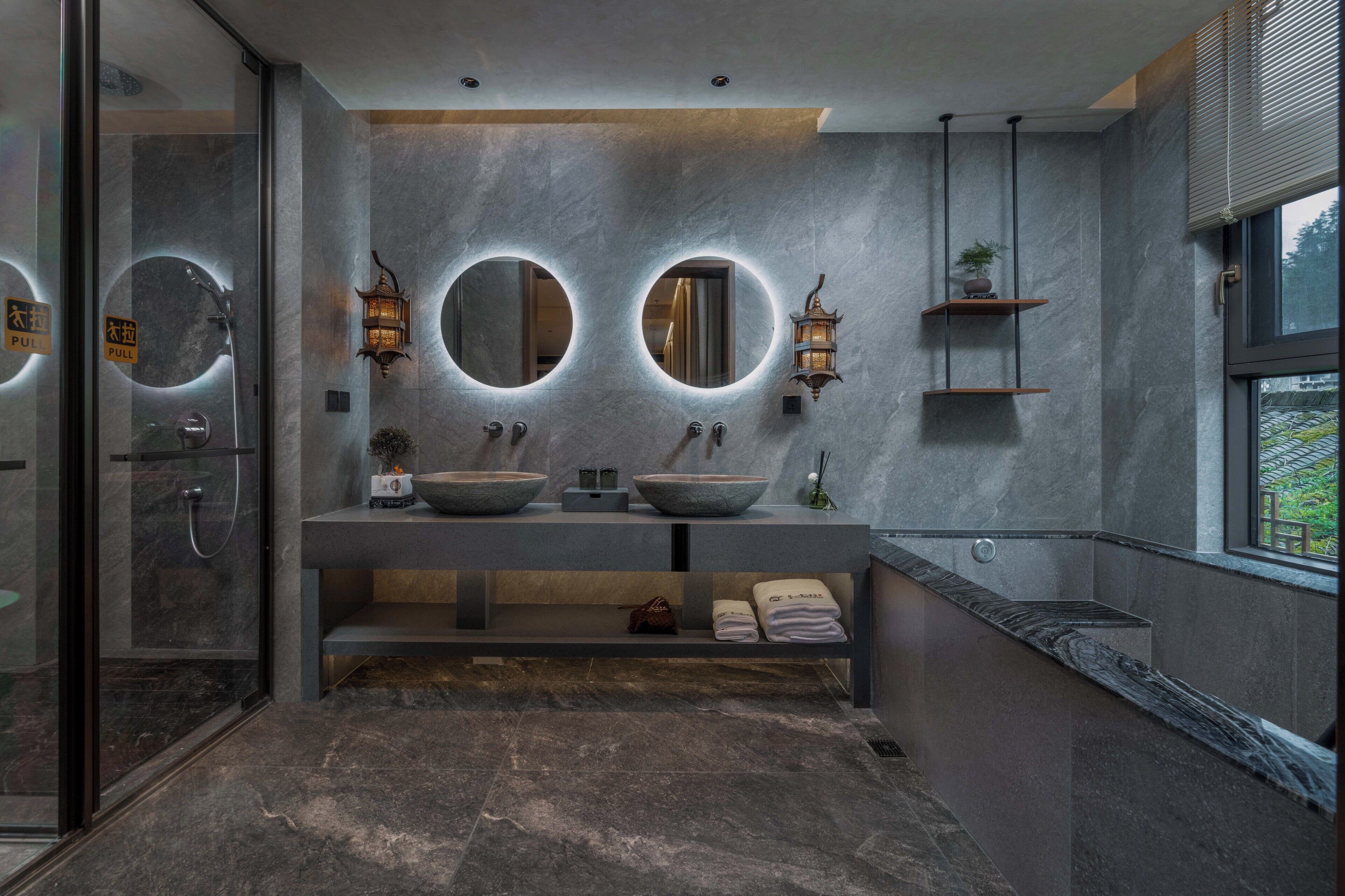 A modern bathroom with two large round mirrors, a glass shower enclosure, and a stone sink.