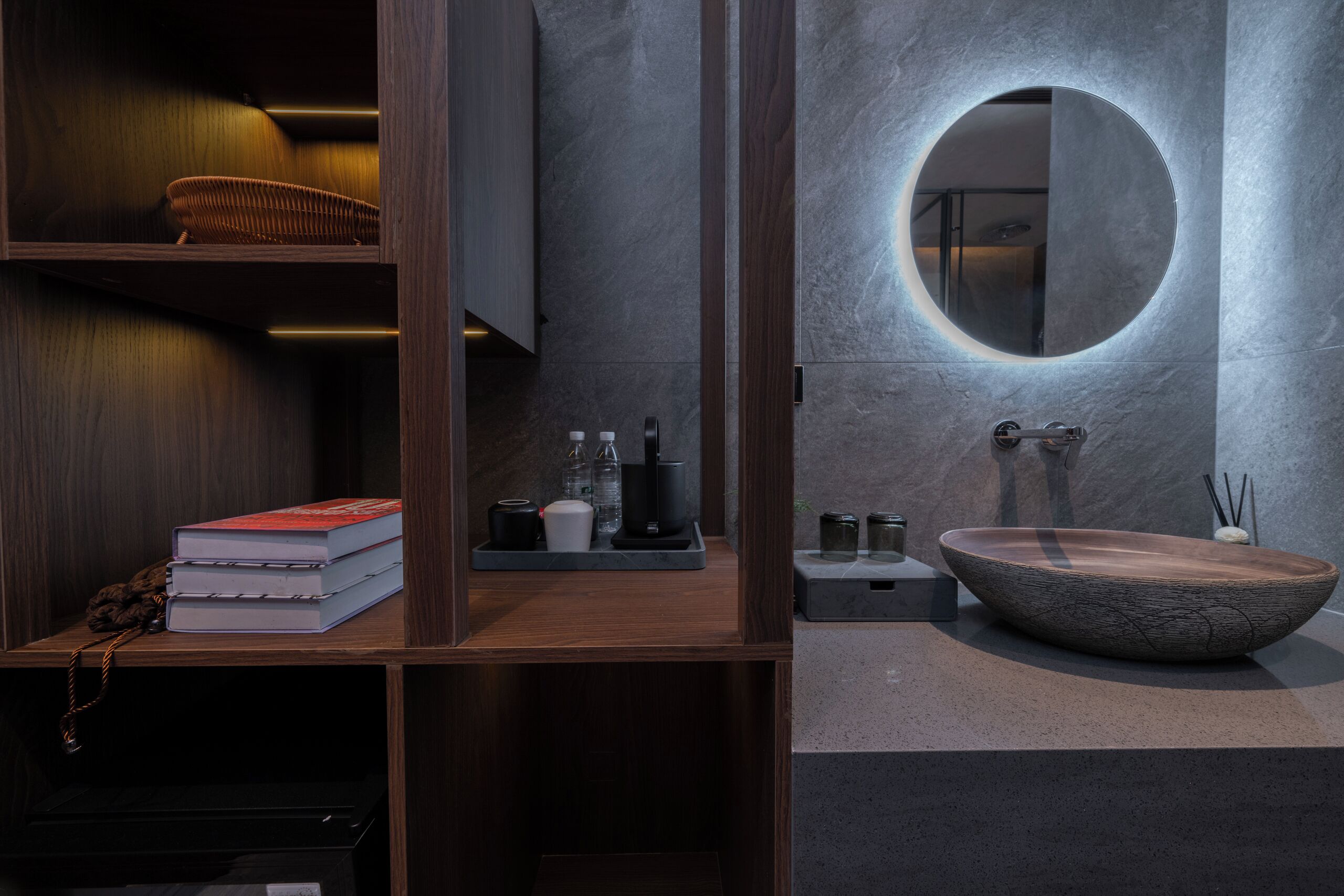A modern bathroom with a round mirror, a wooden shelf with books and toiletries, and a stone sink.