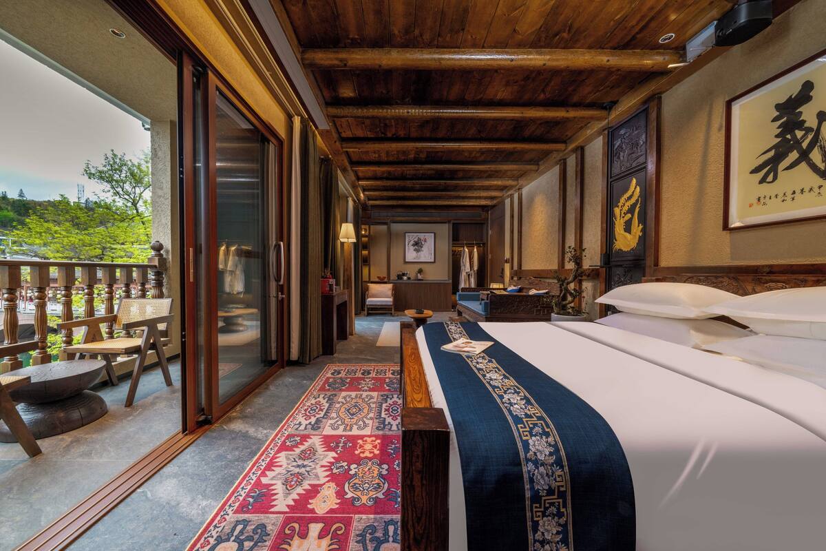 Habitación de hotel con una cama grande, techo de madera, puerta de vidrio corredera a un balcón y una alfombra tradicional.