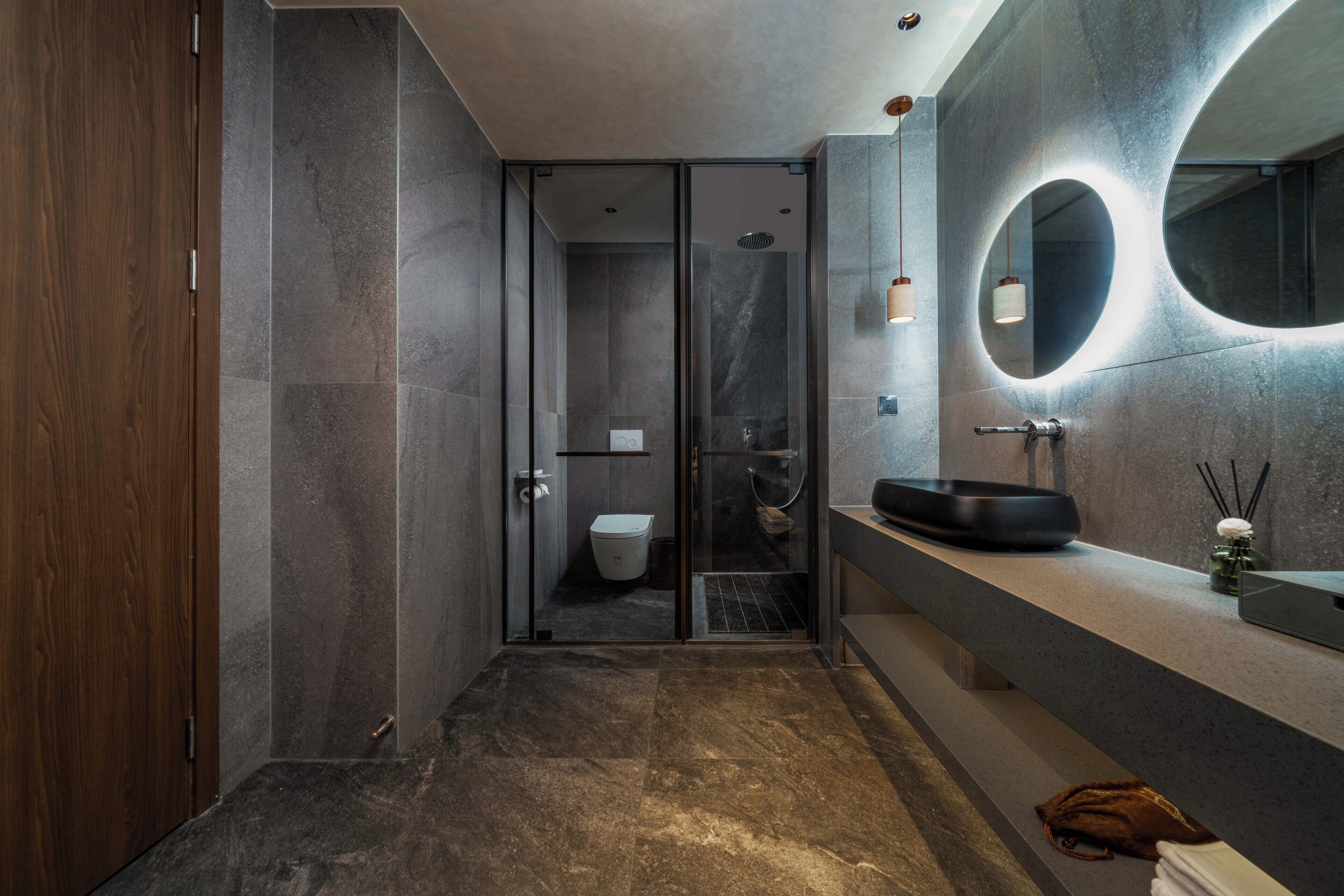 A modern bathroom with a glass shower enclosure, a wooden door, a round mirror, and a black basin.