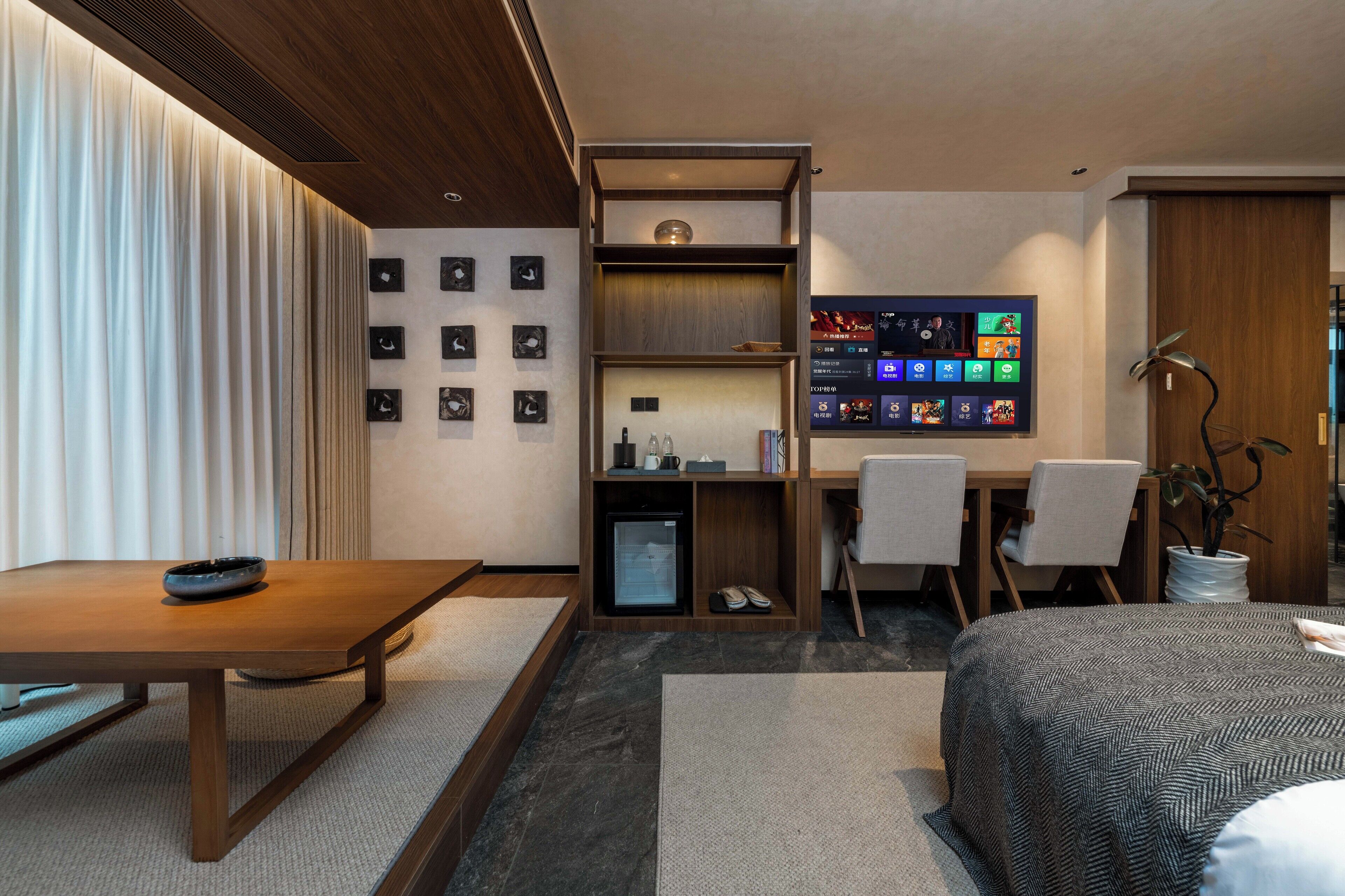 Habitació d'hotel moderna amb escriptori de fusta, llit amb estovatge estampat, petita prestatgeria amb objectes i pantalla digital que mostra contingut.