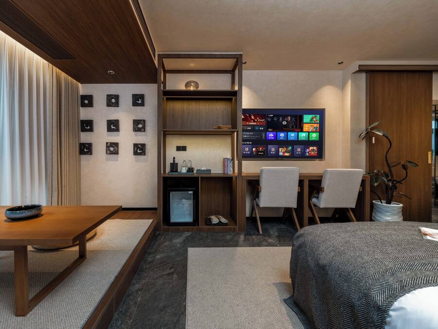 Habitació d'hotel moderna amb escriptori de fusta, llit amb estovatge estampat, petita prestatgeria amb objectes i pantalla digital que mostra contingut.