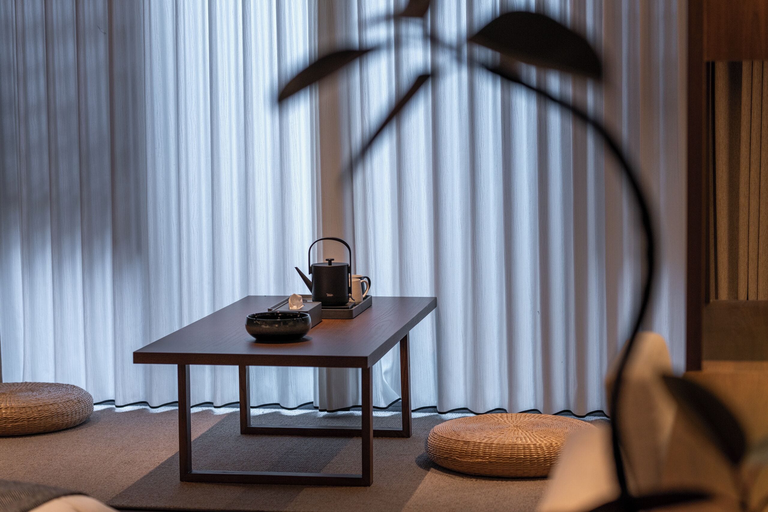 A room with a wooden table, a black kettle, and a teapot on it. There are two woven poufs and a tall floor lamp.
