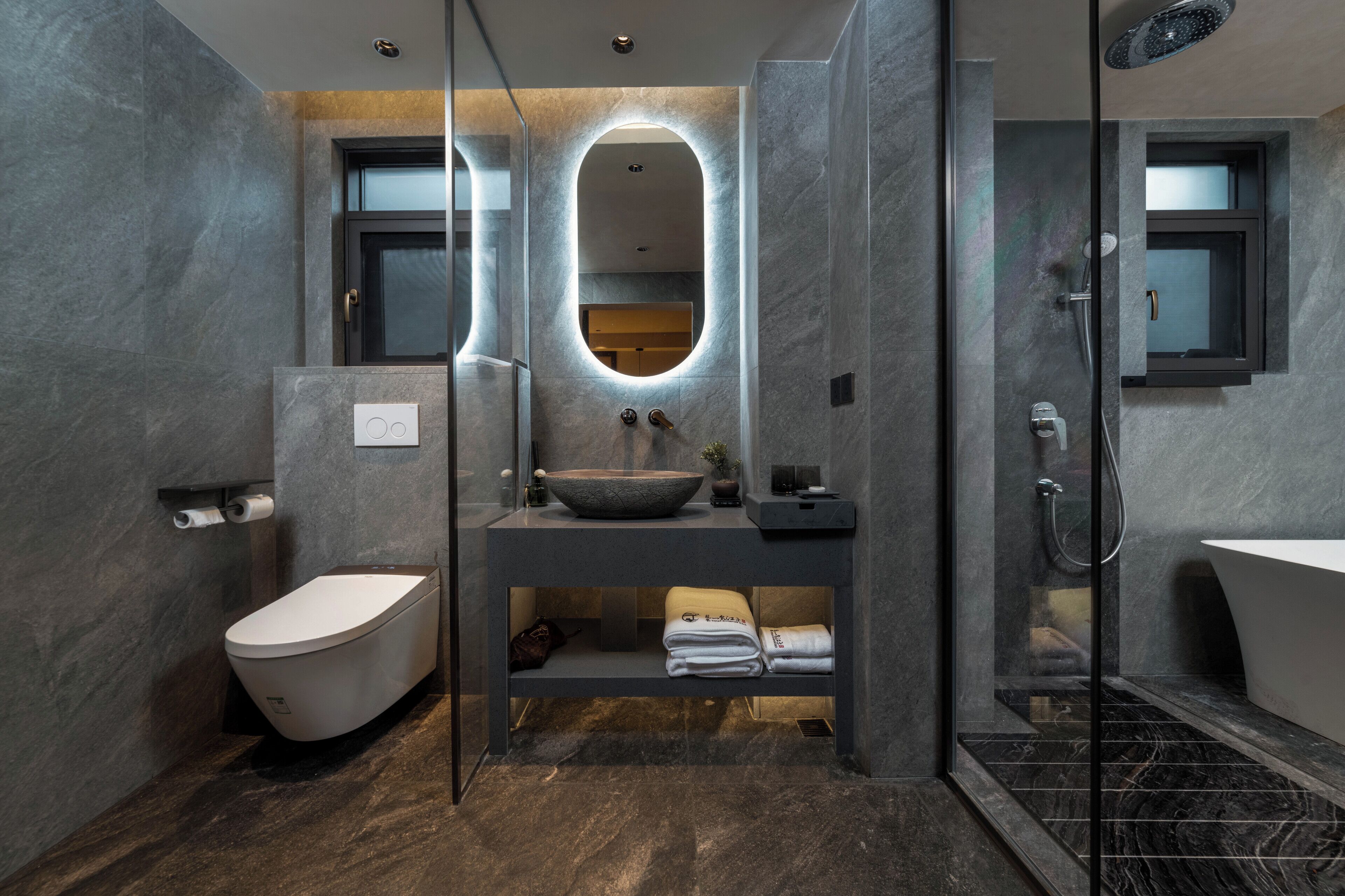 A modern bathroom with a freestanding toilet, a large oval mirror, a built-in sink, and a glass-enclosed shower area.