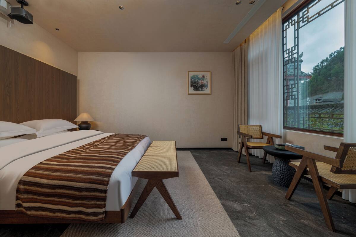 Habitación de hotel con cama, escritorio, silla y ventana con vistas a la vegetación.