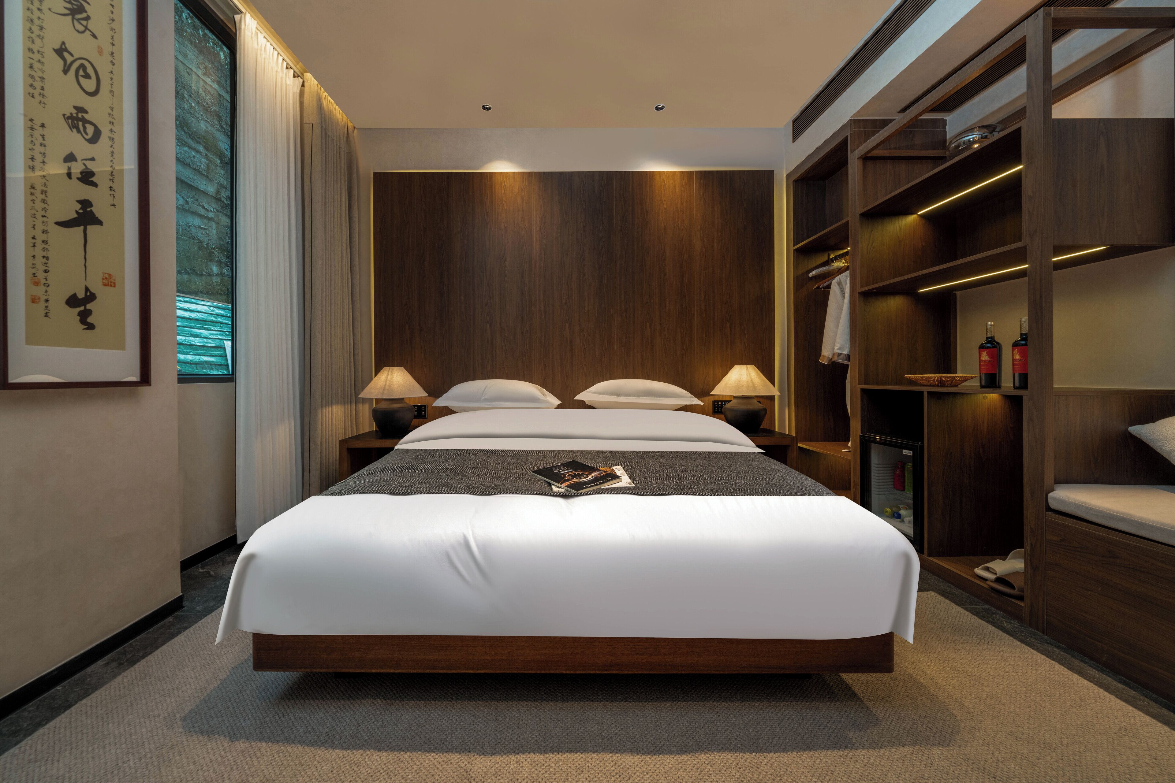 Une chambre d’hôtel moderne avec un grand lit, des lampes de chevet, une tête de lit en bois, une armoire intégrée et une œuvre d’art encadrée au mur.