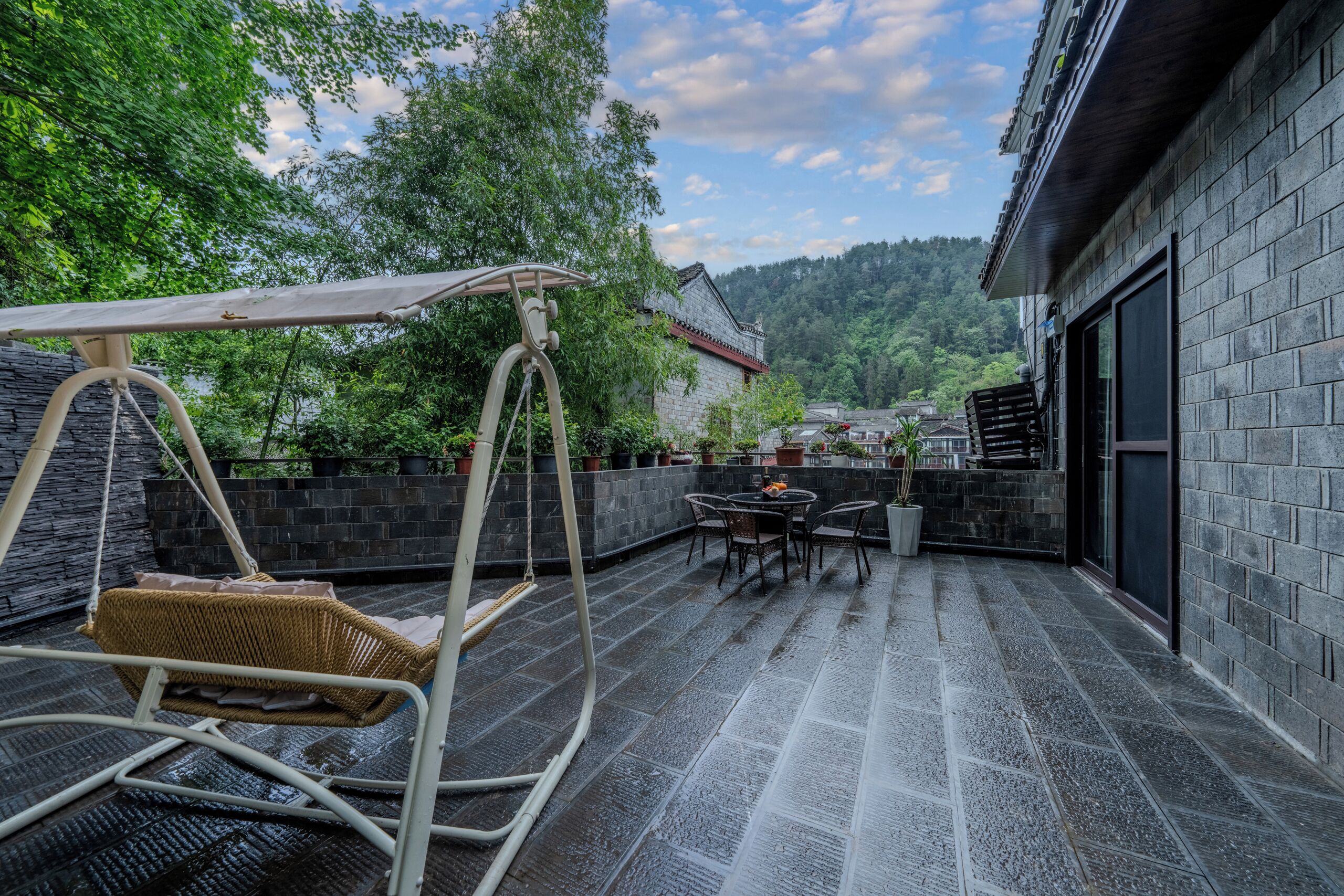 A patio with a swing chair, a table, and chairs, surrounded by potted plants and a stone wall.