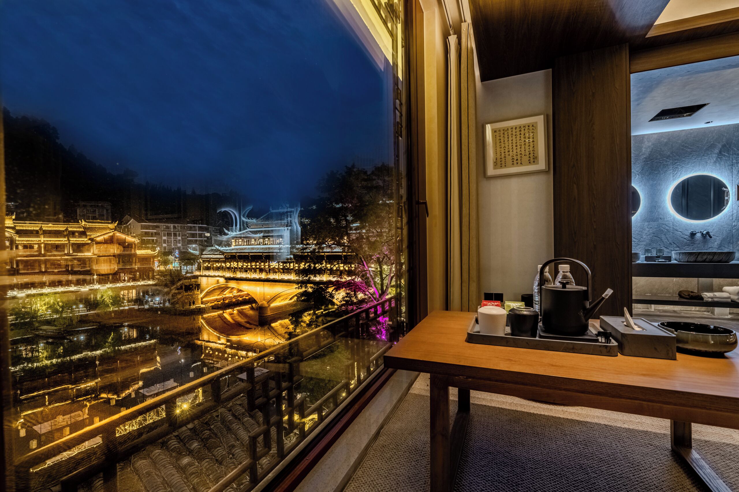 A hotel room with a view of a traditional building at night, a wooden table with tea set, and a modern bathroom with a round mirror.