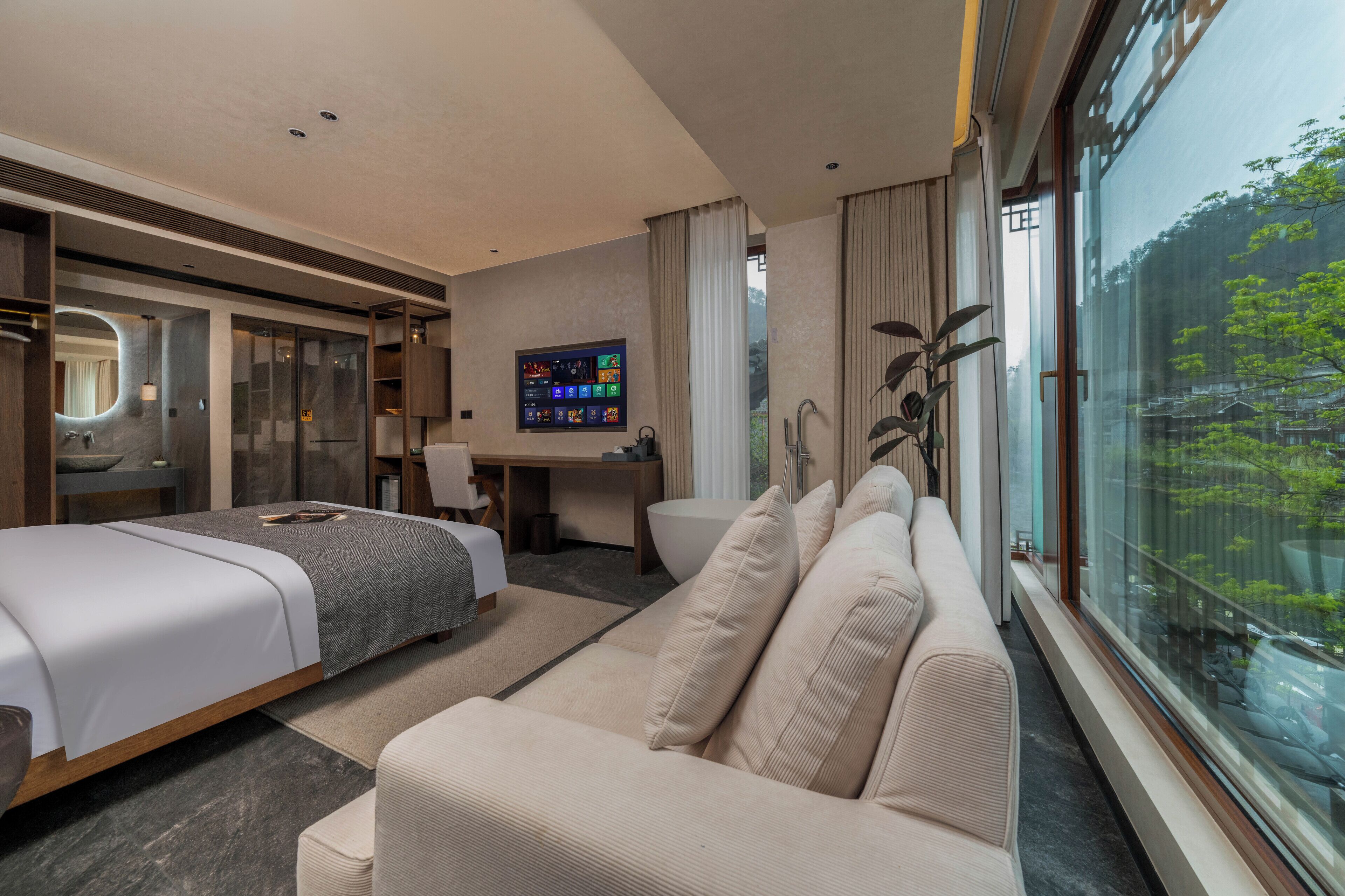 A modern hotel room with a bed, a sofa, a desk, and a large window offering a view of trees.