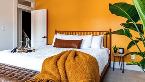 A bedroom with a bed, a white fluffy rug, a brown ottoman, a large plant, and a small wooden side table. - Jungle House Boutique Residences – Downtown Columbus & Short North (Columbus)