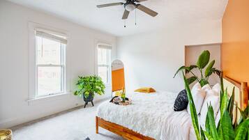 A bedroom with a bed, a ceiling fan, a mirror, a plant, and a window.