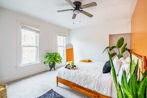 Une chambre à coucher avec un lit, un ventilateur de plafond, un miroir, une plante et une fenêtre.