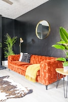 A modern living room with a dark accent wall, a tufted orange sofa, a round mirror, and a small round table with a plant.