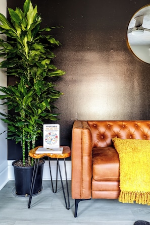 A brown leather sofa with a yellow throw pillow, a potted plant, and a sign on a small stool. - Jungle House Boutique Residences – Downtown Columbus & Short North (Columbus)