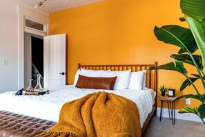 Une chambre à coucher avec un lit, un tapis blanc et moelleux, un pouf marron, une grande plante et une petite table d’appoint en bois.