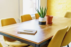 A wooden table with a notebook and pen, surrounded by yellow chairs, and a potted plant.