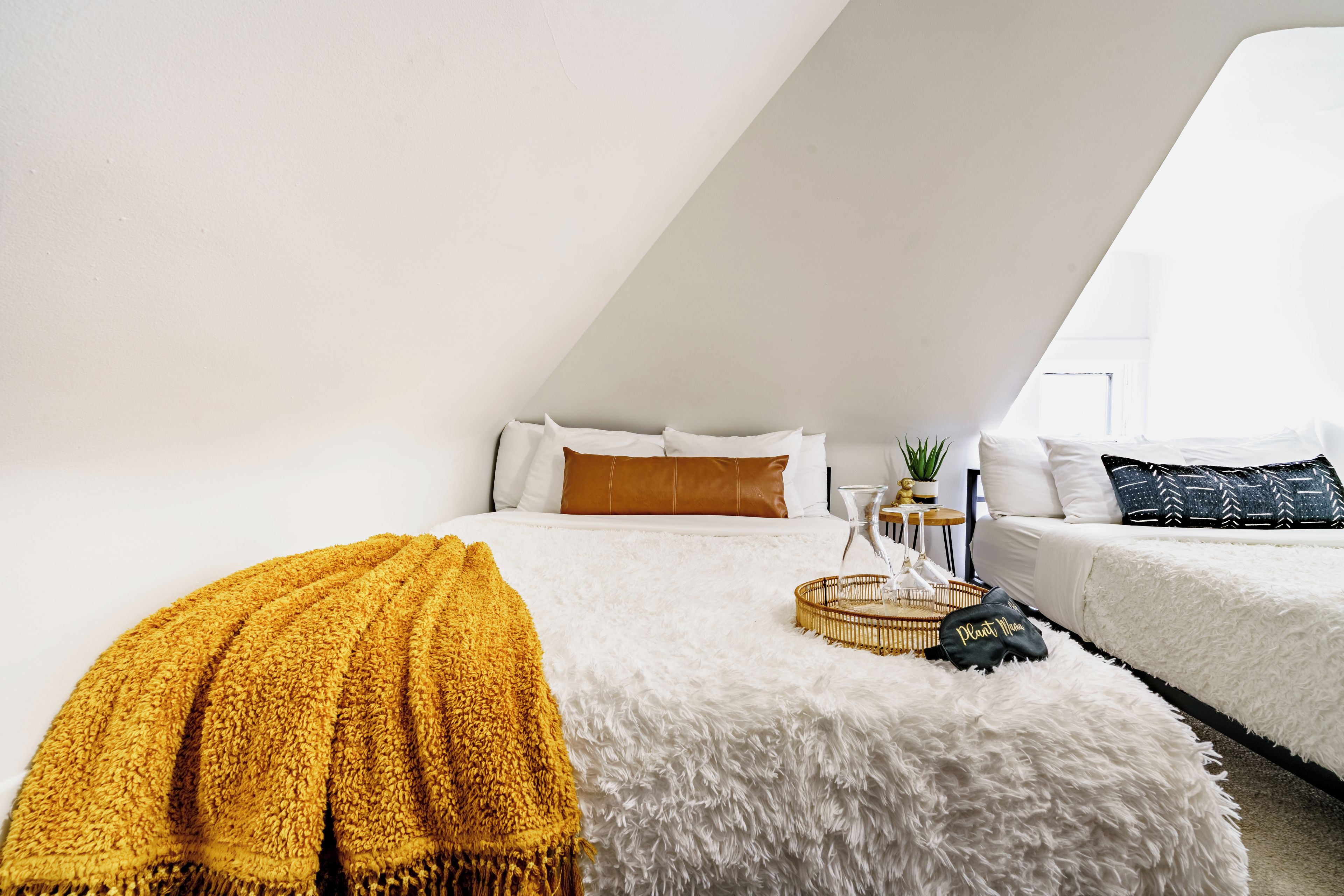 Un dormitorio moderno con techo inclinado, una cama grande con ropa de cama blanca y marrón, una alfombra blanca mullida y una mesita de noche con una planta y un recipiente de vidrio.