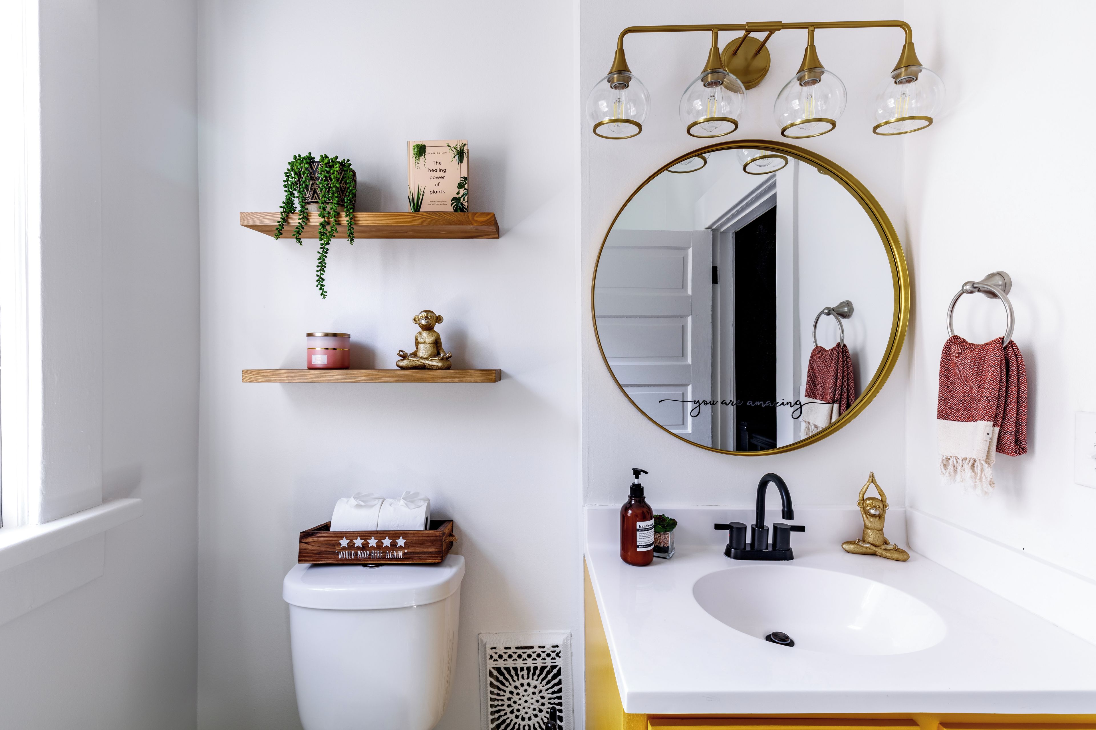 Een badkamer met een wit toilet, een witte wasbak met gouden kraan, een spiegel met gouden lijst, en houten planken met planten en toiletartikelen.