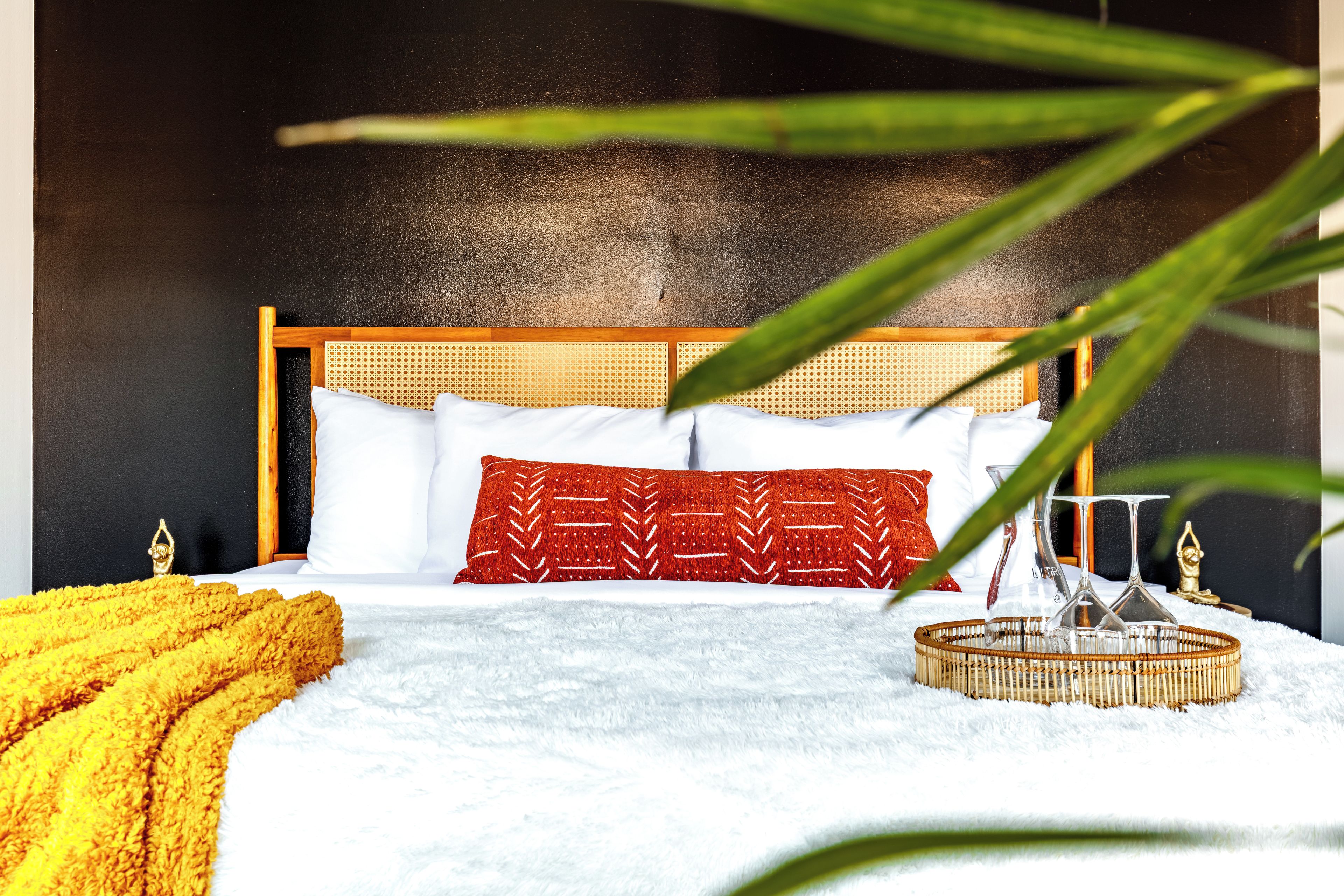 A bed with a wooden headboard, white and red pillows, a yellow throw blanket, and a woven tray with glasses.