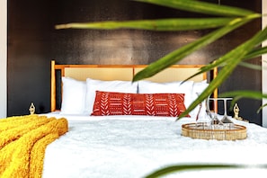 Una cama con cabecera de madera, almohadas blancas y rojas, una manta amarilla y una bandeja tejida con vasos.