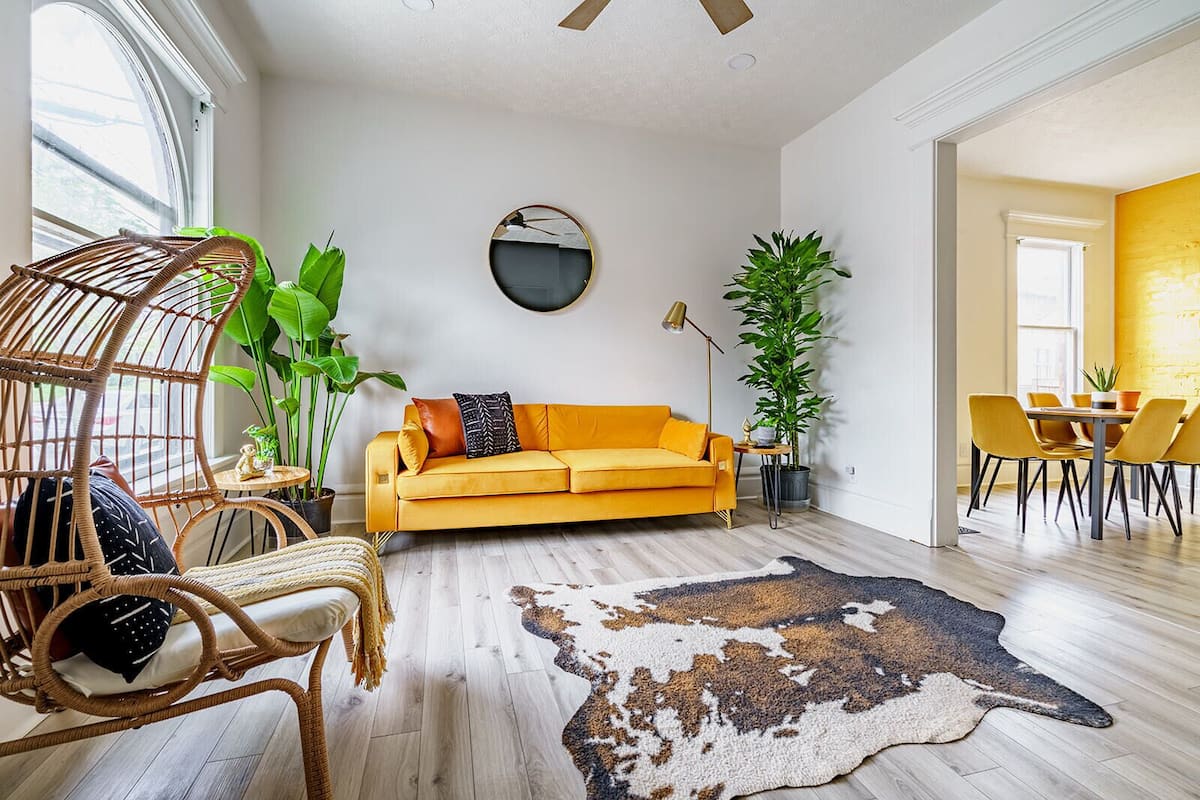 Ein Wohnzimmer mit gelber Couch, Korbsesseln, einem runden Spiegel und einem Essbereich mit gelben Stühlen.