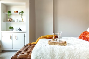 A bedroom with a white bed, a brown leather bench, a white fluffy rug, a white wardrobe, and a shelf with plants and decorative items. - Jungle House Boutique Residences – Downtown Columbus & Short North (Columbus)