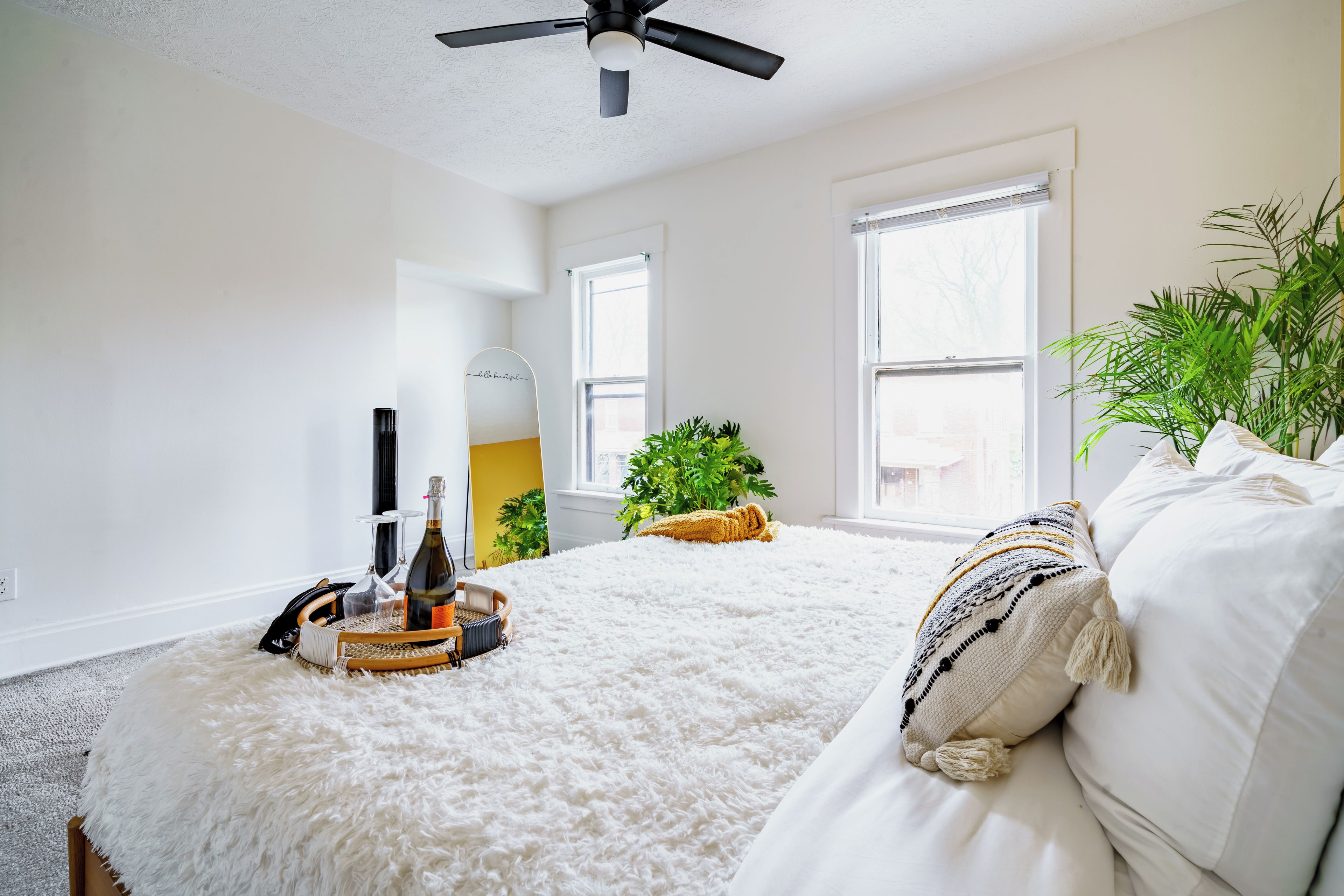 Une chambre à coucher avec un lit, un ventilateur de plafond, deux fenêtres, une plante et une table de chevet sur laquelle se trouvent une bouteille et des verres.