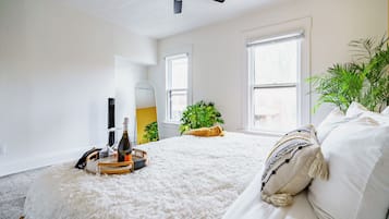 Une chambre à coucher avec un lit, un ventilateur de plafond, deux fenêtres, une plante et une table de chevet sur laquelle se trouvent une bouteille et des verres.