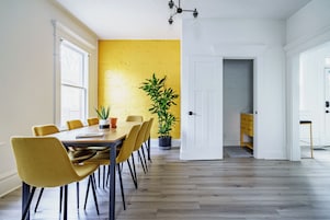 Un comedor con una mesa y sillas de madera, una planta en maceta y un muro de ladrillo amarillo.