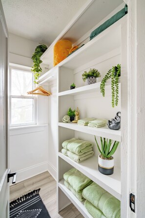 A hallway with white walls, a wooden floor, and a window. Shelves are lined with folded towels, potted plants, and a decorative item. - Jungle House Boutique Residences – Downtown Columbus & Short North (Columbus)