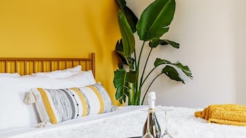 Una cama con una manta blanca y esponjosa, una botella y un vaso sobre una bandeja, una planta y una almohada amarilla.