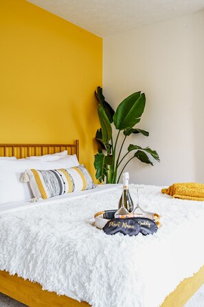 Uma cama com um cobertor branco e fofo, uma garrafa, um copo em um tabuleiro, uma planta e um travesseiro amarelo.