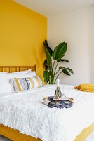 Una cama con una manta blanca y esponjosa, una botella y un vaso sobre una bandeja, una planta y una almohada amarilla.