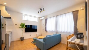 Een moderne woonkamer met een blauwe bank, een eettafel met stoelen, een aan de muur bevestigde televisie en een potplant.