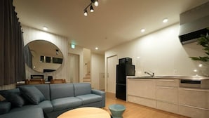 Een moderne woonkamer met een blauwe bank, een ronde houten salontafel, en een keuken met witte kasten en een zwarte koelkast.