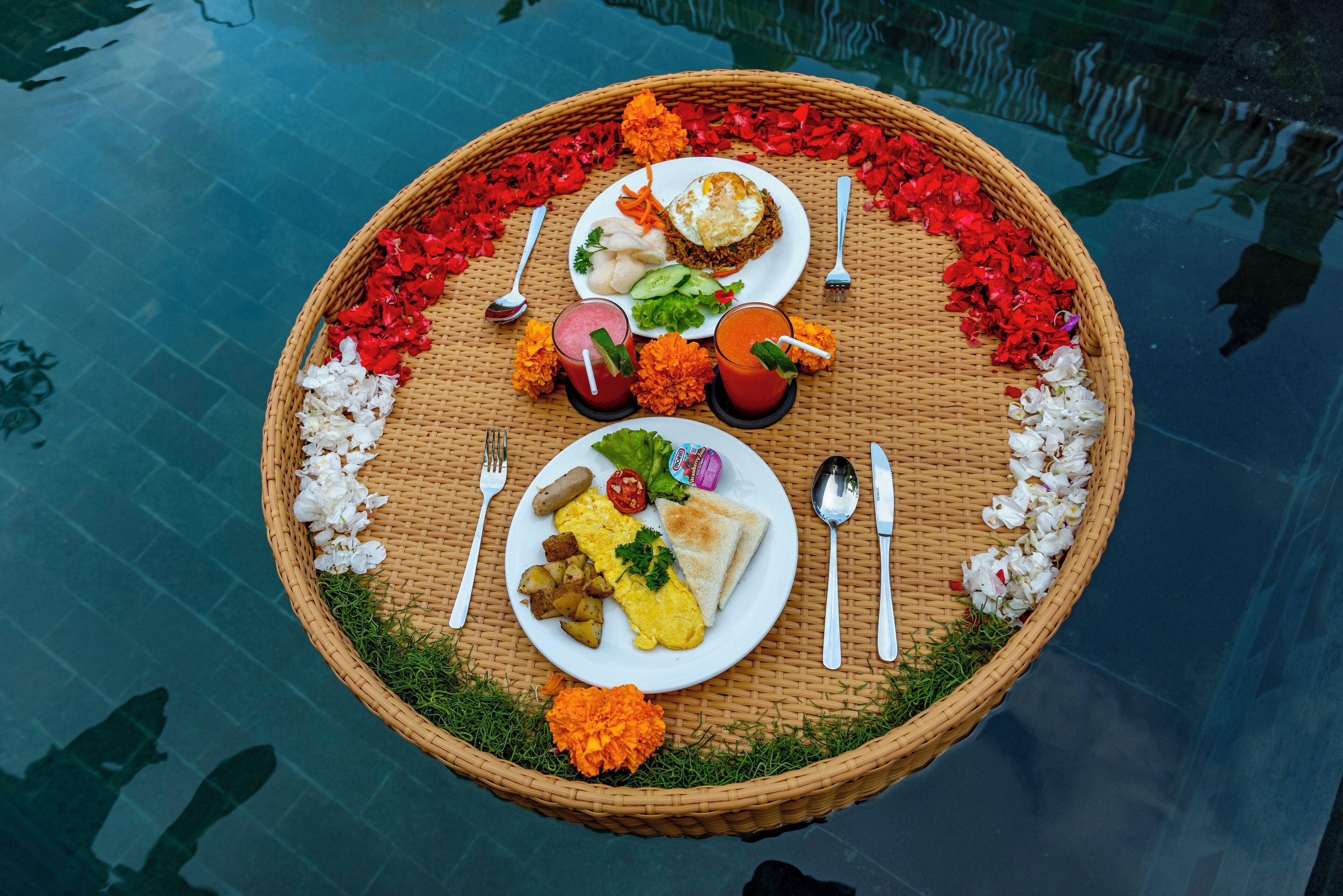 A floating dining setup with a variety of food items and utensils on a woven mat, surrounded by flower petals.